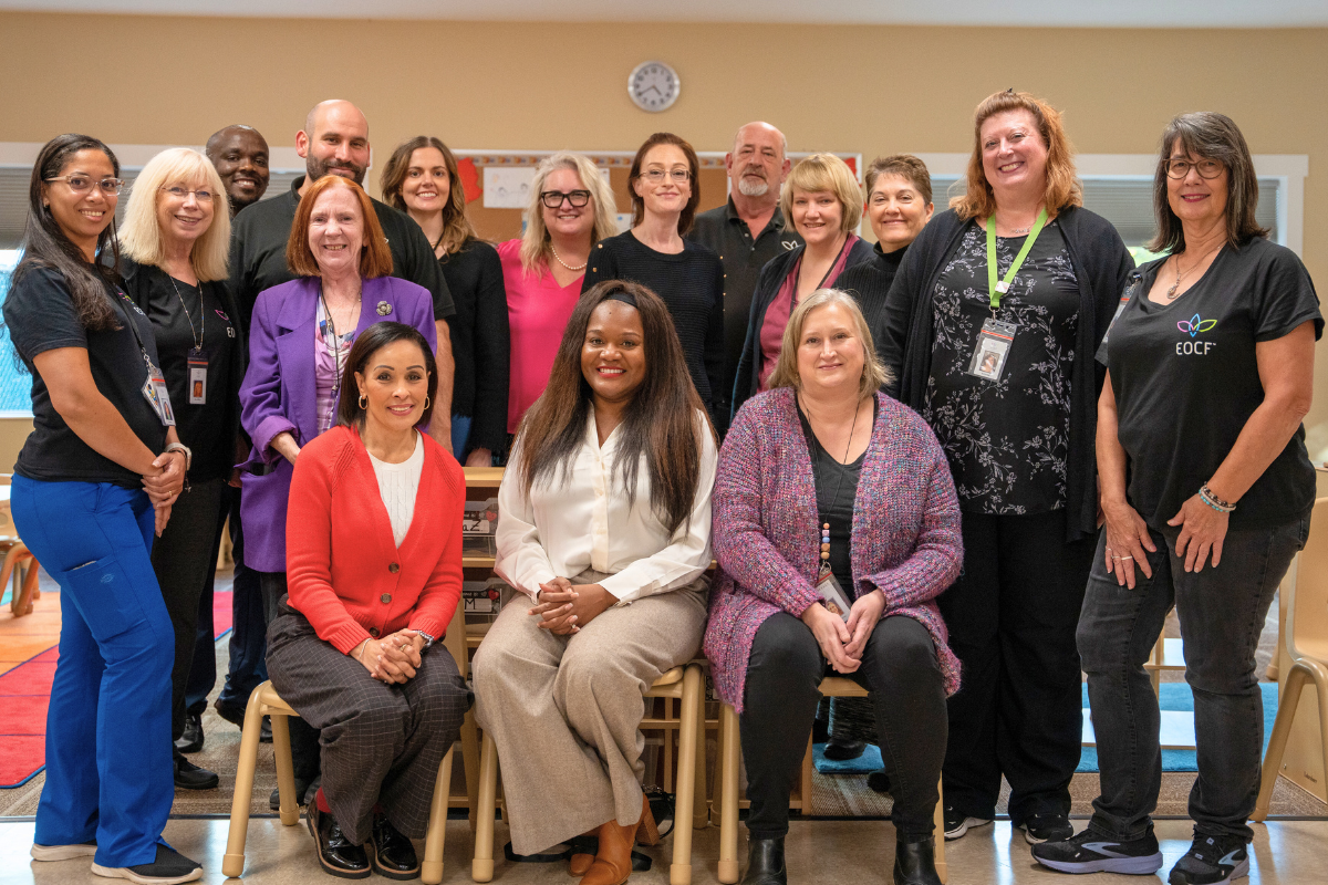 A group of staff members sits in two rows inside a bright, welcoming classroom at the Ruby Bridges Early Learning Center. They are all smiling and dressed in professional attire, exuding a sense of pride and excitement. 